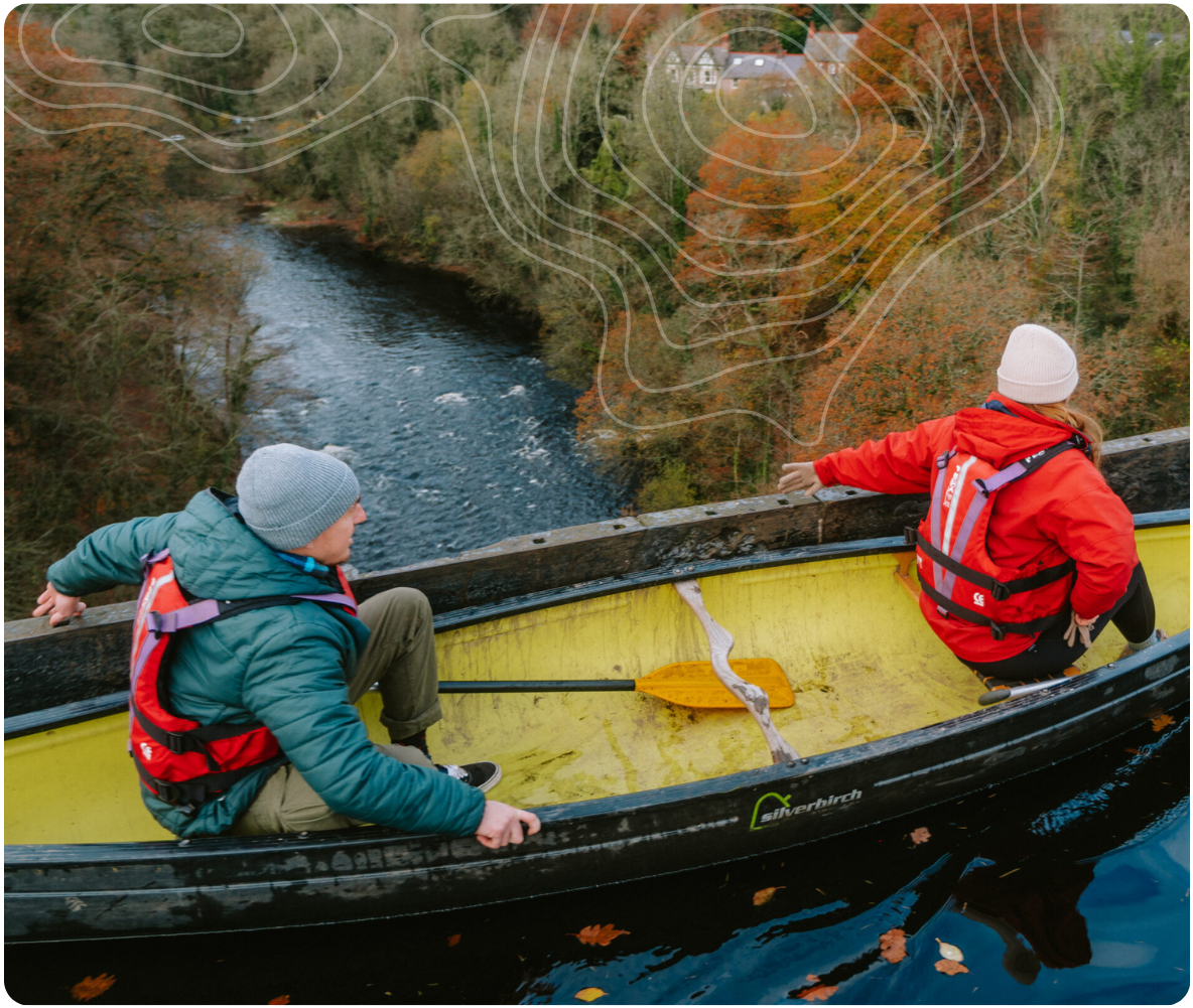 Two people in a canoe on a river surrounded by autumn trees, wearing life jackets and beanies.