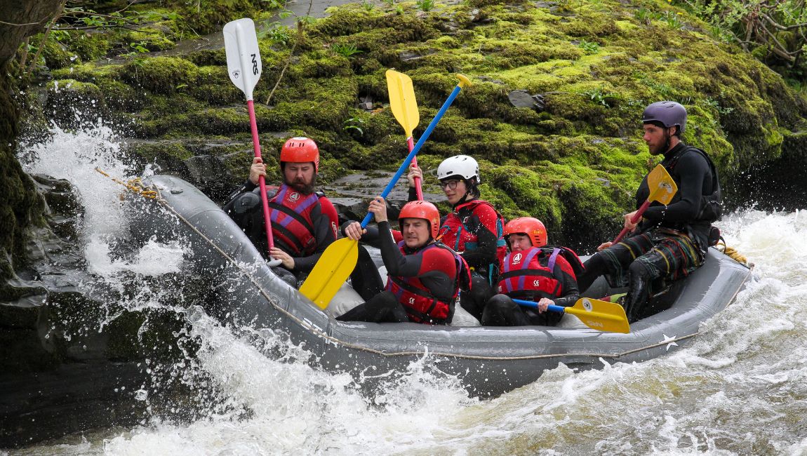 Group rafting on river, wearing helmets and jackets, paddling through rapids near mossy rocks.